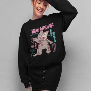 May include: Black sweatshirt featuring a cartoon cat with anatomical details labelled in Japanese. The text reads "猫の解剖学", which translates to "Cat Anatomy", and "あなたの魂を見る目", which translates to "The eye that sees your soul".