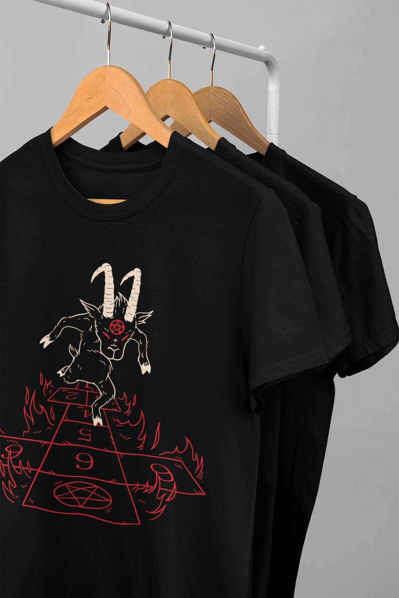 Peut inclure: T-shirt noir avec un motif repr&eacute;sentant un bouc avec des cornes et un pentagramme sur la t&ecirc;te, sautant sur un jeu de marelle enflamm&eacute; avec les chiffres 1, 2, 5, 9 et un pentagramme.