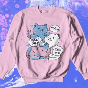 May include: Pink sweatshirt with a graphic of three cartoon cats stacked on top of each other. Each cat has a speech bubble with a different pronoun set: "They/Them", "He/Him", and "She/Her".