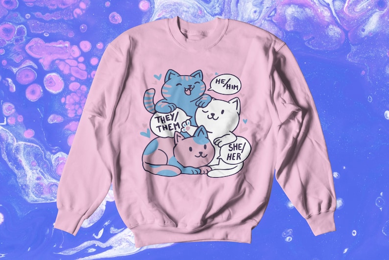 May include: Pink sweatshirt with a graphic of three cartoon cats stacked on top of each other. Each cat has a speech bubble with a different pronoun set: "They/Them", "He/Him", and "She/Her".