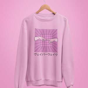 May include: A light pink sweatshirt with a graphic design featuring two hands reaching towards each other within a pink and black grid. Japanese text is below the graphic. The sweatshirt is hanging on a wooden hanger.