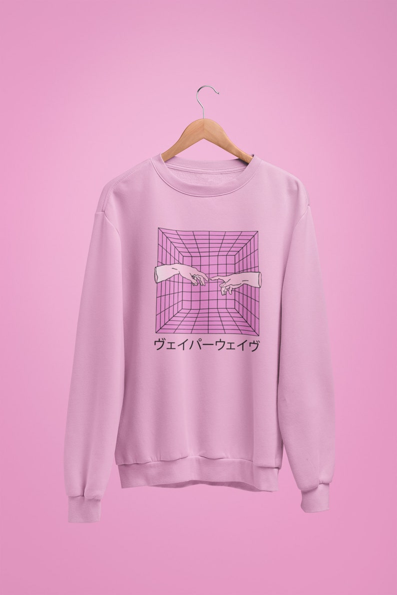 May include: A light pink sweatshirt with a graphic design featuring two hands reaching towards each other within a pink and black grid. Japanese text is below the graphic. The sweatshirt is hanging on a wooden hanger.