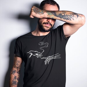May include: Black t-shirt with a white graphic of two hands reaching towards each other, one human and one skeletal, holding a cigarette with smoke rising from it. The text "The Creation of Adam" is not visible in the image.