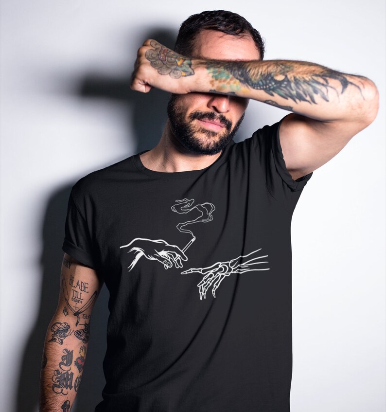 May include: Black t-shirt with a white graphic of two hands reaching towards each other, one human and one skeletal, holding a cigarette with smoke rising from it. The text "The Creation of Adam" is not visible in the image.