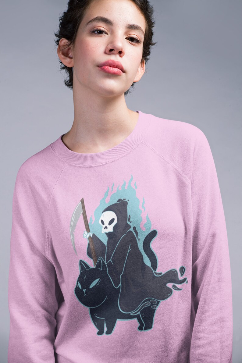 K&ouml;nnte beinhalten: Ein rosa Sweatshirt mit einem Grafikdruck des Sensenmanns, der auf einer schwarzen Katze reitet. Der Sensenmann h&auml;lt eine Sense und ist von blauen Flammen umgeben.