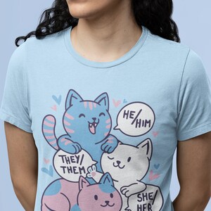 May include: A light blue t-shirt with a graphic of three cartoon cats. The cats are stacked on top of each other. The top cat is blue and white, the middle cat is white, and the bottom cat is pink and blue. Each cat has a speech bubble with text. The top cat's speech bubble says "He/Him", the middle cat's speech bubble says "They/Them", and the bottom cat's speech bubble says "She/Her".