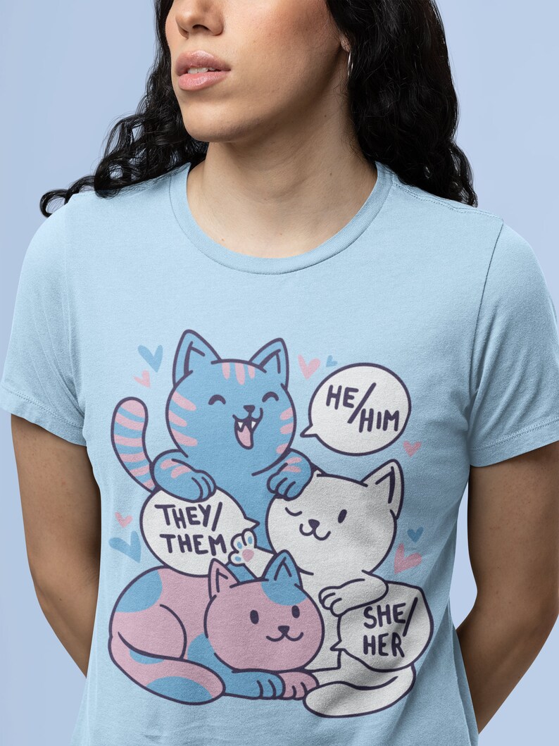 May include: A light blue t-shirt with a graphic of three cartoon cats. The cats are stacked on top of each other. The top cat is blue and white, the middle cat is white, and the bottom cat is pink and blue. Each cat has a speech bubble with text. The top cat's speech bubble says "He/Him", the middle cat's speech bubble says "They/Them", and the bottom cat's speech bubble says "She/Her".