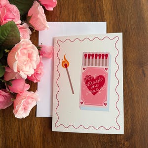 May include: A white greeting card with a red scalloped border featuring a graphic of a matchbox and a lit match. The matchbox is pink with a red heart and the text "love struck the match" written in white script. A white envelope is included.