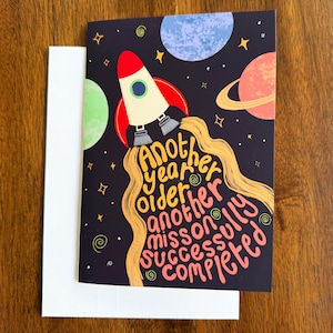 May include: A greeting card featuring a dark space background with planets, stars, and a rocket ship. The rocket emits yellow exhaust trails that form the text: Another year older, another mission successfully completed. A plain white envelope is included.