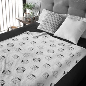 Puede incluir: Una manta de forro polar blanca con un patrón de dados blanco y negro. La manta está sobre una cama con sábanas negras y dos almohadas blancas.