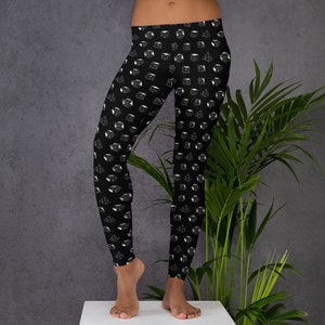 May include: Black leggings featuring a repeating pattern of white dice. The leggings are close-fitting and extend to the ankles. The dice are in various shapes, including cubes and pyramids. The background is a dark grey.
