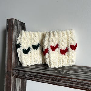 Peut inclure: Deux manchons de tasse en crochet crème avec des motifs en chevrons. L'un a des chevrons vert foncé, l'autre des chevrons rouges. Les manchons sont présentés sur une étagère en bois rustique.