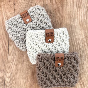 Peut inclure: Trois chauffe-tasses en crochet beige, blanc et marron avec des lanières en cuir qui disent "Coffee Cup Cozy".