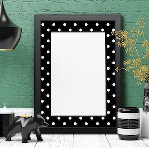 May include: A black picture frame with a white polka dot border sits on a white surface. The frame is empty and ready for a photo or artwork. There are black candles, a black bear figurine, and a black and white striped vase on the surface.