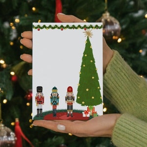 May include: A white card with a green Christmas tree and three nutcracker figurines. The card has a red and gold garland at the top and a red base with presents.