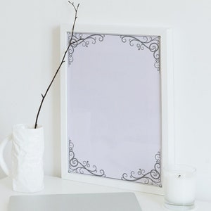 May include: A white picture frame with a decorative border of gray swirls. The frame is on a white table with a white vase and a twig inside and a white candle on the right.