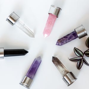 May include: Overhead view of six crystal wands with silver metal caps, arranged in a circle. The crystals are clear, pink, purple, black, and smoky quartz. A sprig of green and purple leaves is visible.