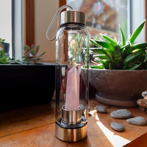 Puede incluir: Una botella de agua de vidrio transparente con tapa y asa de acero inoxidable. Un cristal rosa está dentro de la botella. La botella está sobre una superficie de madera.