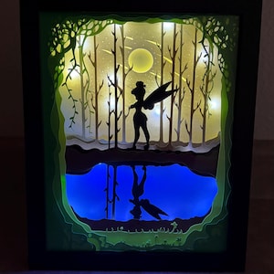 May include: A rectangular black-framed shadow box featuring a multi-layered paper cutout scene. A silhouette of a winged figure stands in a forest at night with a yellow moon, illuminated by internal lights, reflecting into a blue water surface.