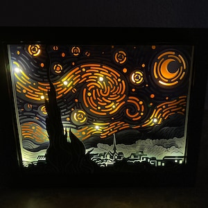 May include: A black framed light-up art piece depicting Vincent van Gogh's Starry Night. The artwork features a swirling sky with a crescent moon and stars, a dark silhouette of a village, and a bright yellow and orange color scheme.