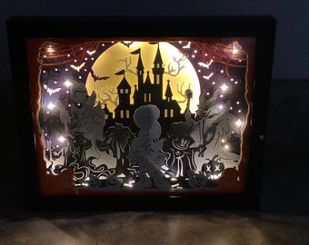 May include: A Halloween-themed shadow box with a spooky castle, bats, and various characters. The layered design creates a 3D effect, illuminated by small lights. The color scheme includes orange, black, and yellow, evoking a festive atmosphere.