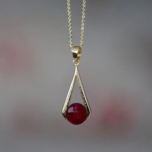 Op de afbeelding: Een goudkleurige ketting met een hanger. De hanger heeft een gefacetteerde, dieprode edelsteen, gezet in een goudkleurig, open driehoekig frame. De ketting is delicaat. Een sieraad.