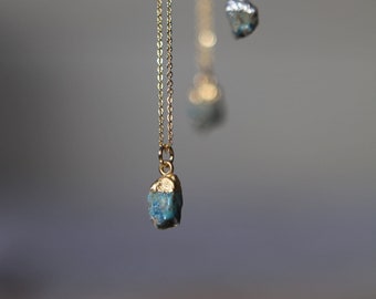 May include: Close-up of three necklaces with small, irregular blue-green gemstone pendants. One necklace is gold-toned, while the others are silver-toned. The chains are delicate, and the background is a soft gray.