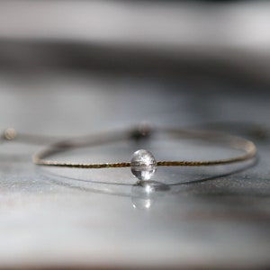 Pode incluir: Uma pulseira delicada com uma conta transparente em forma oval centrada em uma fina faixa dourada. A faixa tem uma superfície texturizada. A pulseira está colocada sobre um fundo neutro desfocado.