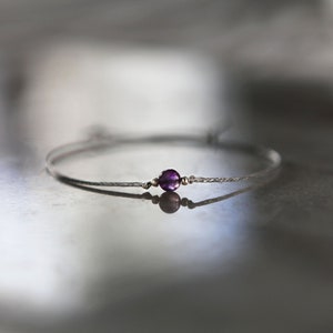May include: A delicate silver bracelet featuring a small, round, deep purple amethyst gemstone. The bracelet has a thin, textured band and is displayed on a reflective surface, creating a soft, diffused reflection.