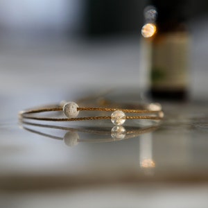 May include: A delicate gold-toned bracelet with clear and textured white beads. The bracelet is lying on a reflective surface, with a blurred background that includes a bottle. The bracelet is a piece of jewelry.