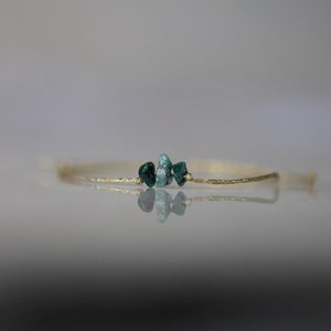 Op de afbeelding: Een delicate goudkleurige armband met drie kleine, onregelmatig gevormde, teal-groene edelstenen. De armband heeft een dunne, getextureerde band en wordt gepresenteerd tegen een zachte, vage achtergrond.