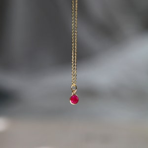 Op de afbeelding: Een delicate gouden ketting met een kleine, ronde, robijnrode edelsteen hanger. De hanger hangt aan een fijne, gouden ketting. De achtergrond is een zachte, vage grijze en witte gradiënt.