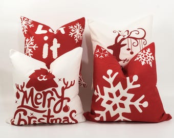 May include: Four decorative Christmas throw pillows. Two pillows are white with red designs, including snowflakes and the text "Let it". The other two pillows are red with white designs, including a reindeer and the text "Merry Christmas".