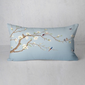 May include: A rectangular decorative pillow with a light blue background. The pillow features a detailed illustration of a flowering tree branch with white blossoms and several colorful birds. The branches are brown, and the birds have red, blue, and gray feathers.