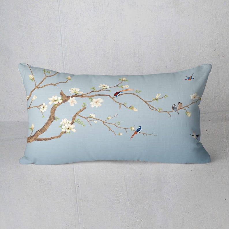 May include: A rectangular decorative pillow with a light blue background. The pillow features a detailed illustration of a flowering tree branch with white blossoms and several colorful birds. The branches are brown, and the birds have red, blue, and gray feathers.