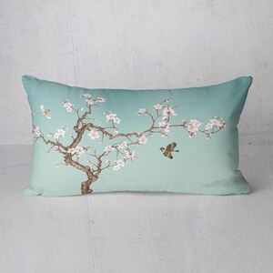 May include: Rectangular decorative pillow with a serene design. The pillow features a light teal background with a watercolor illustration of a flowering tree branch, small birds, and soft pink blossoms. The design evokes a sense of tranquility.