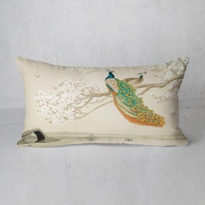 May include: Rectangular decorative pillow featuring a watercolor painting of two peacocks perched on a flowering tree branch. The birds have blue, green, and gold plumage. The background includes a bridge and a small boat.