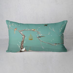 May include: Rectangular decorative pillow with a teal background featuring a tree with birds and a birdcage. The tree branches have white blossoms. The birds are in shades of brown, orange, and gray. The birdcage is gold.