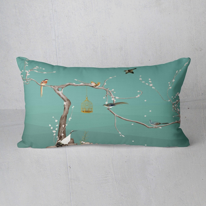 May include: Rectangular decorative pillow with a teal background featuring a tree with birds and a birdcage. The tree branches have white blossoms. The birds are in shades of brown, orange, and gray. The birdcage is gold.