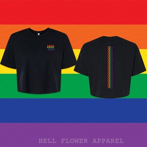 Pu&ograve; includere: Una t-shirt nera corta con un piccolo quadrato a scacchi arcobaleno sul petto sinistro e una lunga striscia verticale a scacchi arcobaleno centrata sul retro. Il capo presenta maniche corte e un girocollo, mostrato su uno sfondo arcobaleno.