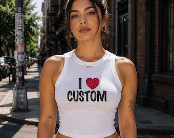 May include: A white sleeveless crop top featuring the text I, a red glitter heart, and the word CUSTOM in black lettering. The shirt includes the text HELL FLOWER APPAREL printed in the upper right corner. The design is centered on the chest.
