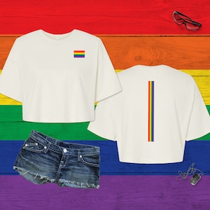 Pu&ograve; includere: Un top corto color crema a maniche corte con una piccola bandiera arcobaleno rettangolare sul petto e una lunga striscia verticale arcobaleno sul retro, abbinato a pantaloncini di jeans blu consumati e occhiali da vista neri.