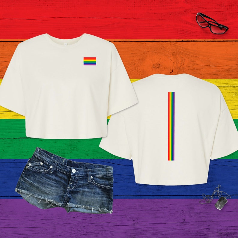 Pu&ograve; includere: Un top corto color crema a maniche corte con una piccola bandiera arcobaleno rettangolare sul petto e una lunga striscia verticale arcobaleno sul retro, abbinato a pantaloncini di jeans blu consumati e occhiali da vista neri.