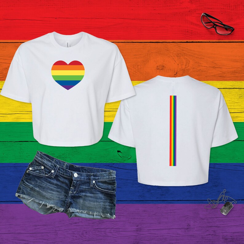 Pu&ograve; includere: Un top corto bianco a maniche corte con un cuore dai colori dell'arcobaleno sul davanti e una striscia verticale arcobaleno sul retro, abbinato a pantaloncini di jeans blu, occhiali con montatura nera e una collana con piastrina militare.