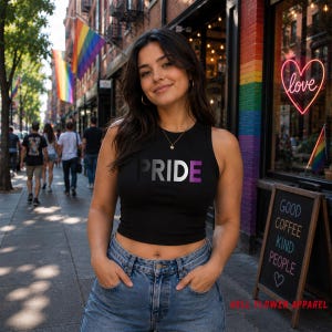 May include: Black crop top with the word "PRIDE" in a gradient of colors, with the "A" in black and the "E" in purple. The model is wearing blue jeans and standing on a sidewalk. Rainbow flags are visible in the background.