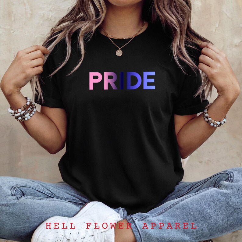 Pode incluir: Uma camiseta preta com a palavra "PRIDE" impressa em rosa, azul e branco. As letras est&atilde;o em um degrad&ecirc; de cores.