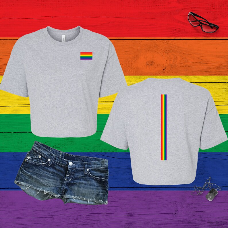 Pu&ograve; includere: Un top corto grigio chiaro a maniche corte con una piccola bandiera arcobaleno orizzontale sul petto sinistro e una striscia arcobaleno verticale sul retro, abbinato a pantaloncini di jeans blu con orlo sfrangiato e una collana con piastrina militare.