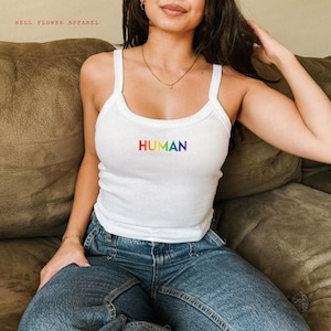 Può includere: Un top bianco a spalline con la parola "HUMAN" stampata in colori arcobaleno. La modella indossa jeans blu.