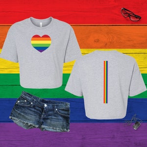 Pu&ograve; includere: Una t-shirt corta grigio chiaro con un cuore dai colori dell'arcobaleno sul davanti e una striscia verticale arcobaleno sul retro, abbinata a pantaloncini di jeans blu, occhiali con montatura nera e una collana con piastrina color argento.