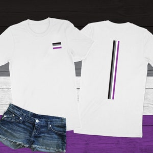 May include: White t-shirt with a black, gray, and purple stripe on the back and a small black, gray, and purple flag on the front. The shirt is shown on a wooden background.